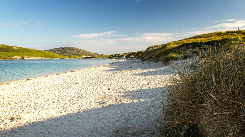 Ein weißer Sandstrand, der sich bis zu grünen Hügeln hinzieht. Die Sonne scheint, der Himmel ist blau.