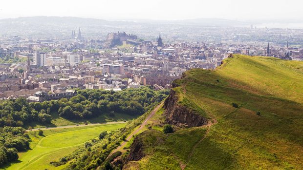 Der Blick vom Arthur's Seat geht über die Klippen der Solisbury Crags im Vordergrund. Dahinter ist die Stadt Edinburgh zu sehen, die Burg sticht prominent heraus.