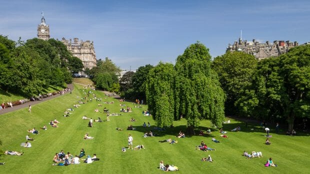 Die Princess Street Gardens ist ein Park. Auf einer grünen Wiese liegen und sitzen Menschen in der Sonne. Im Hintergrund ist das Balmoral Hotel zu sehen. - Foto: Martin Goldmann