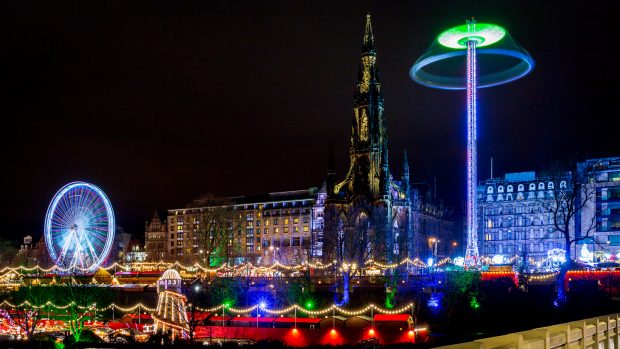 Der Edinburgh Weihnachtsmarkt in den Princes Street Gardens bei Nacht. Das Riesenrad links ist blau erleuchtet, überall sind Lichtergirlanden, das Scott Monument überragt alles.