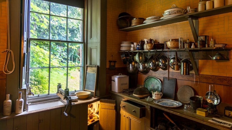 Eine alte Küche mit Holzregalen und alten Küchengeräten. Ein Fenster erhellt die Küche. 