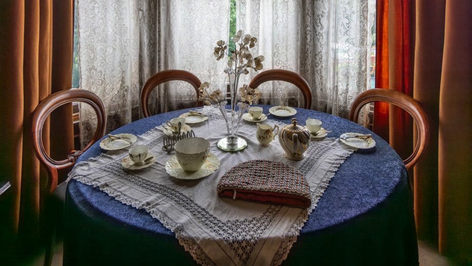 Ein runder, mit Teegeschirr gedeckter Tisch mit Decke und vier alten Stühlen vor einem Fenster
