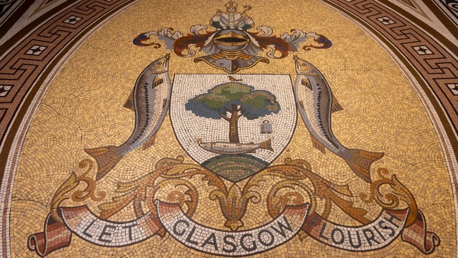 Ein Stadtwappen mit zwei Fischen an der Seite und einem Ritterhelm sowie einem Bischof darüber. Zentrales Motiv ist der Baum mit einem Fisch darunter, der einen goldenen Ring im Maul trägt. Darunter der Spruch "Let Glasgow flourish"