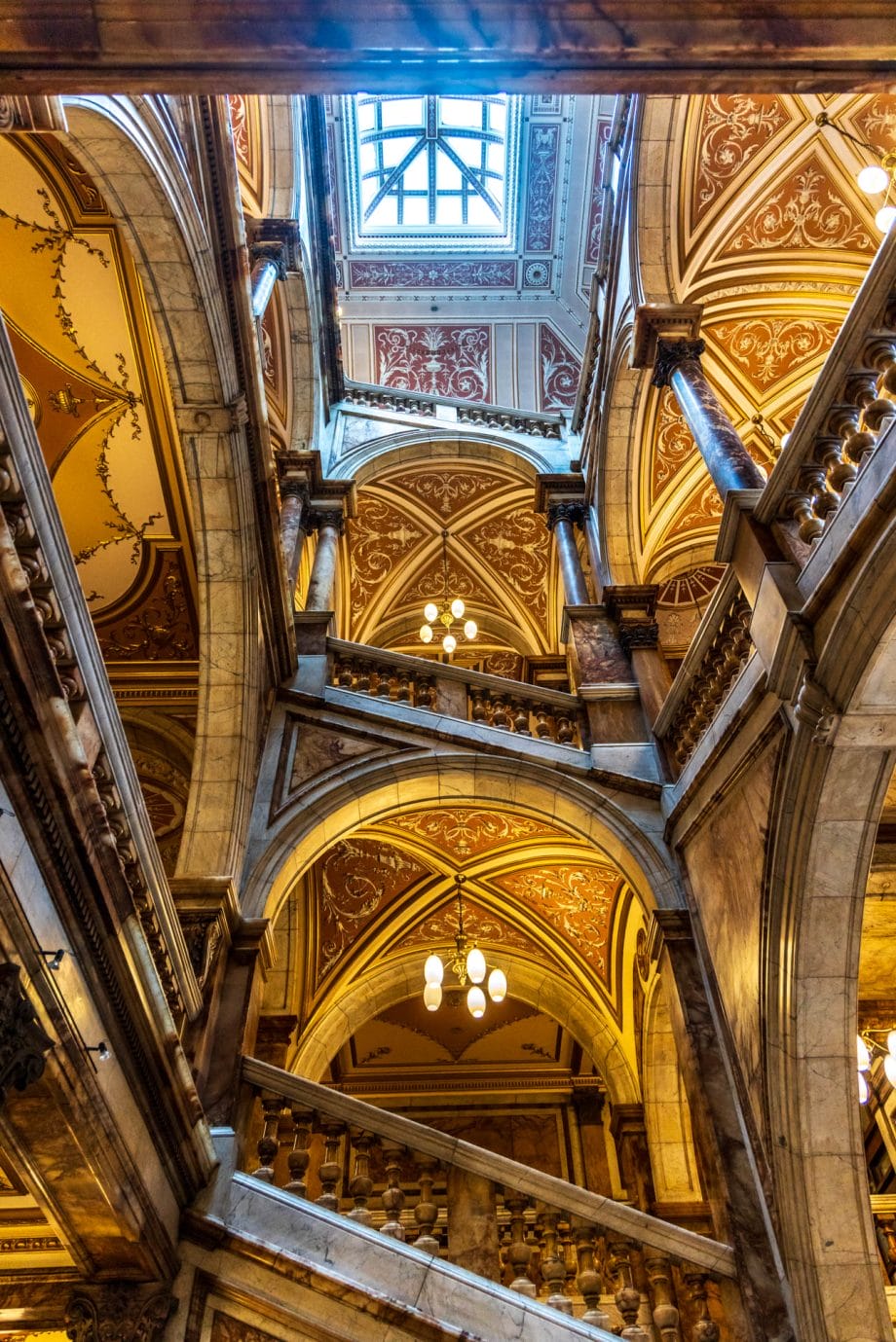 Blick nach oben in ein dreistöckiges Treppenhaus mit Oberlicht. Mit Bögen, Säulen und viel Marmor. 