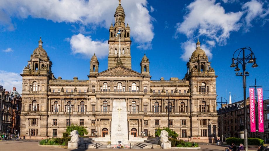 Ein großes graues Gebäude am Ende des Platzes George Square in Glasgow, mit einem großen Turm in der Mitte, zwei kleinen Turmspitzen an den Ecken und einer Fassade, die an die klassische Antike erinnert.