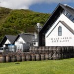 Ein großes, weißes und modernes Gebäude mit Spitzdach. Davor viele Holzfässer. Dahinter blauer Himmel und ein grüner Hang.