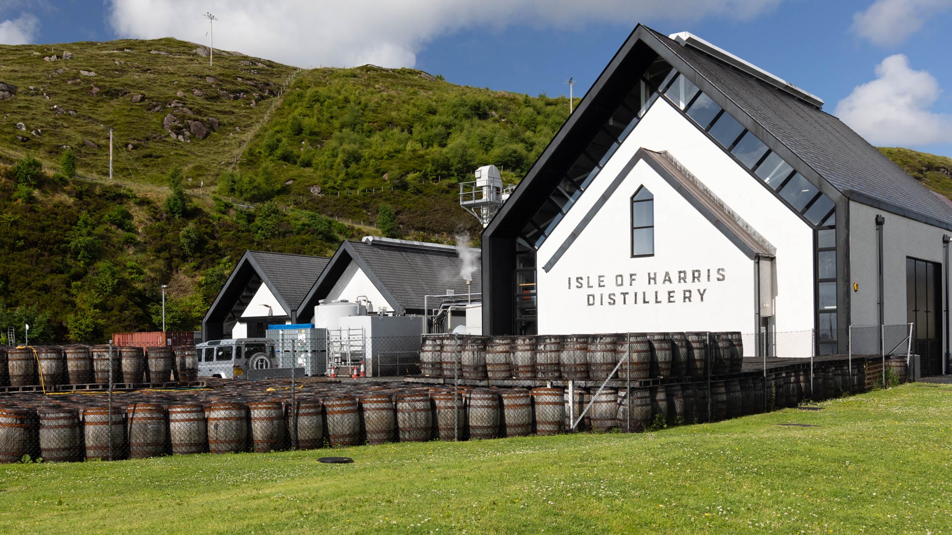 Ein großes, weißes und modernes Gebäude mit Spitzdach. Davor viele Holzfässer. Dahinter blauer Himmel und ein grüner Hang.