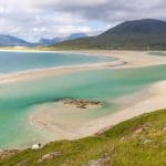 Blick von der Straße auf einen Sandstrand mit grünblauem Wasser und Bergen im Hintergrund auf der Isle of Harris