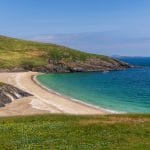 Ein beiger Sandstrand zieht sich an einer Bucht entlang, eingerahmt von Felsen und grünen Hügeln