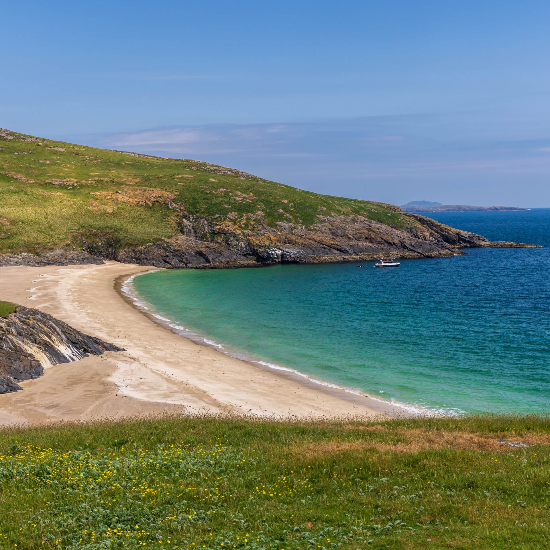 Ein beiger Sandstrand zieht sich an einer Bucht entlang, eingerahmt von Felsen und grünen Hügeln