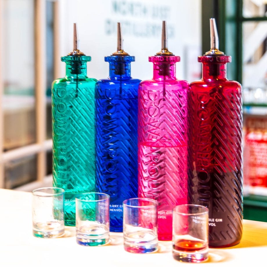 Vier Flaschen in vier verschiedenen Farben stehen hinter vier kleinen durchsichtigen Trinkgläsern