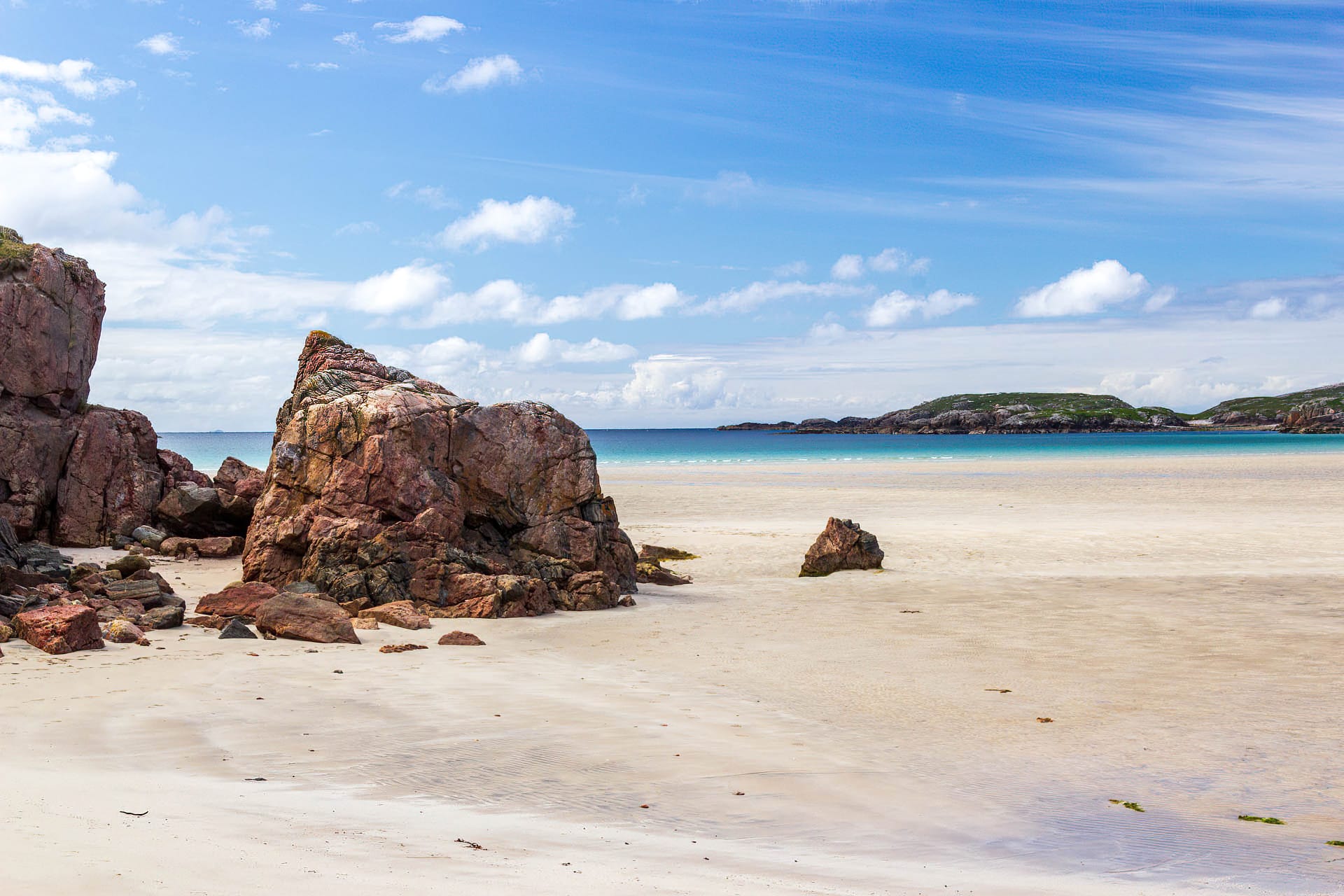 Ein rötlicher Fels ragt aus dem Sand eines weiten Strandes. Im Hintergrund ist das Meer zu sehen.