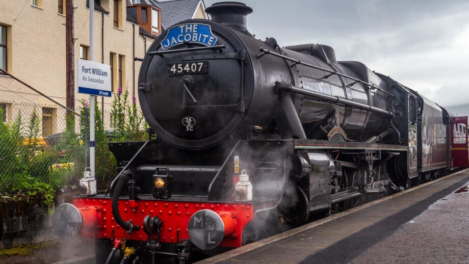 Eine alte, schwarze Dampflokomotive an einem Bahnsteig. Sie steht unter Dampf. Vorne prangt ein blaues Schild mit "The Jacobite" darauf.