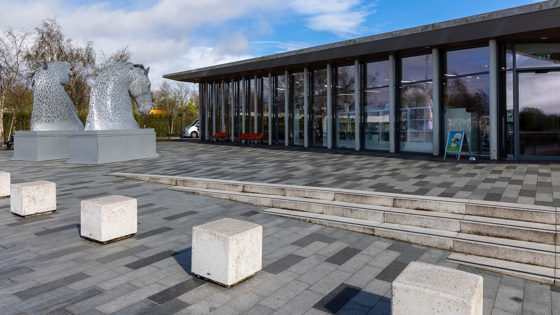 In einem flaches Haus mit Glasfronten befindet sich ein Café. Davor stehen die kleinen Statuen der Kelpies.