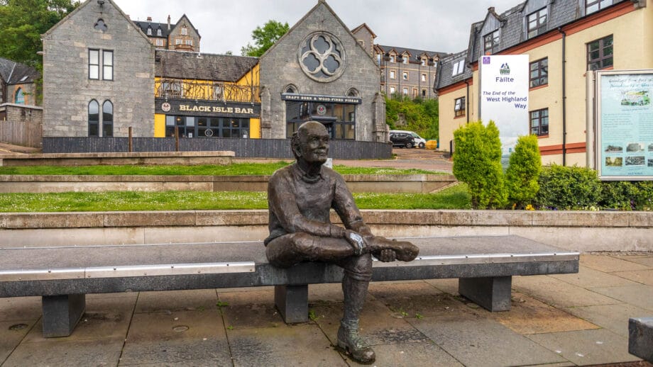 Die Bronze-Statue eines Wanderers sitzt auf einer Bank. Ein Schild dahinter markiert das Ende des West Highland Ways.