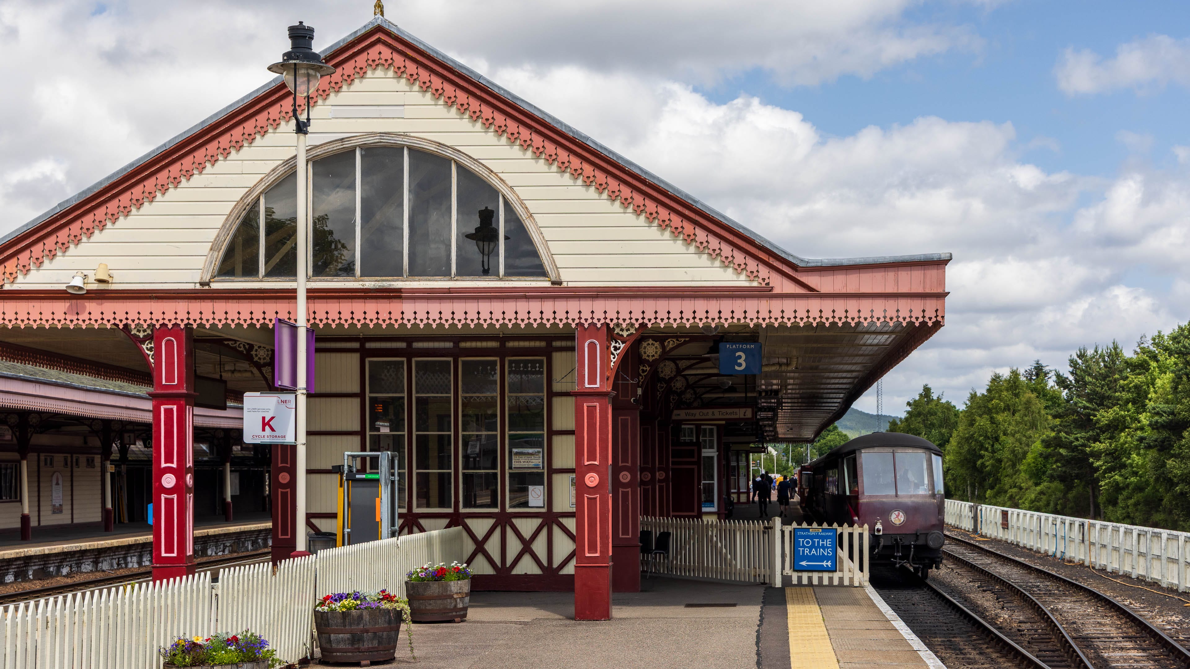 Das Ende des Zuges ist auf einem Gleis zu sehen, daneben erhebt sich das rot-weiße viktorianische Bahnhofsgebäude von Aviemore.