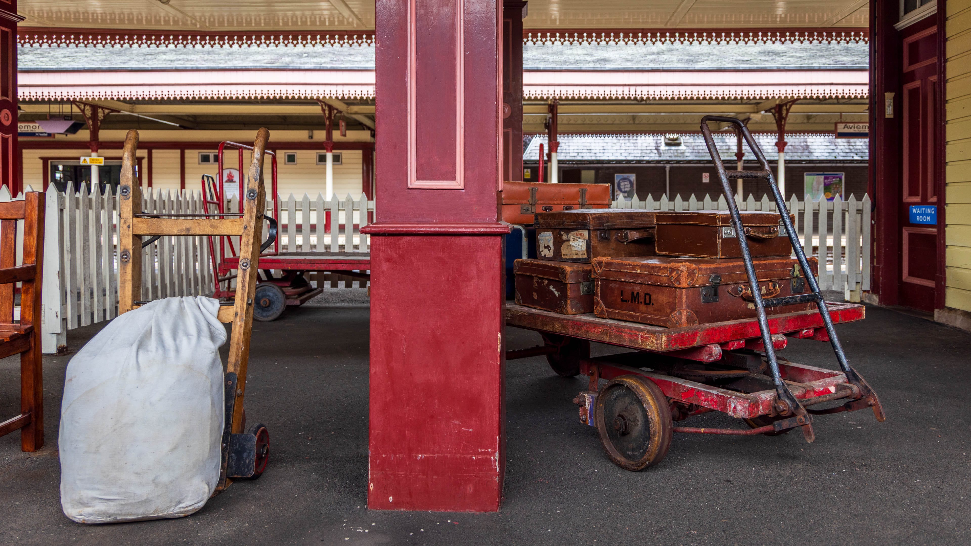Um eine Säule des Bahnhofsgebäudes von Aviemore steht ein historischer Gepäckwagen mit Koffern darauf und eine alte Sackkarre.
